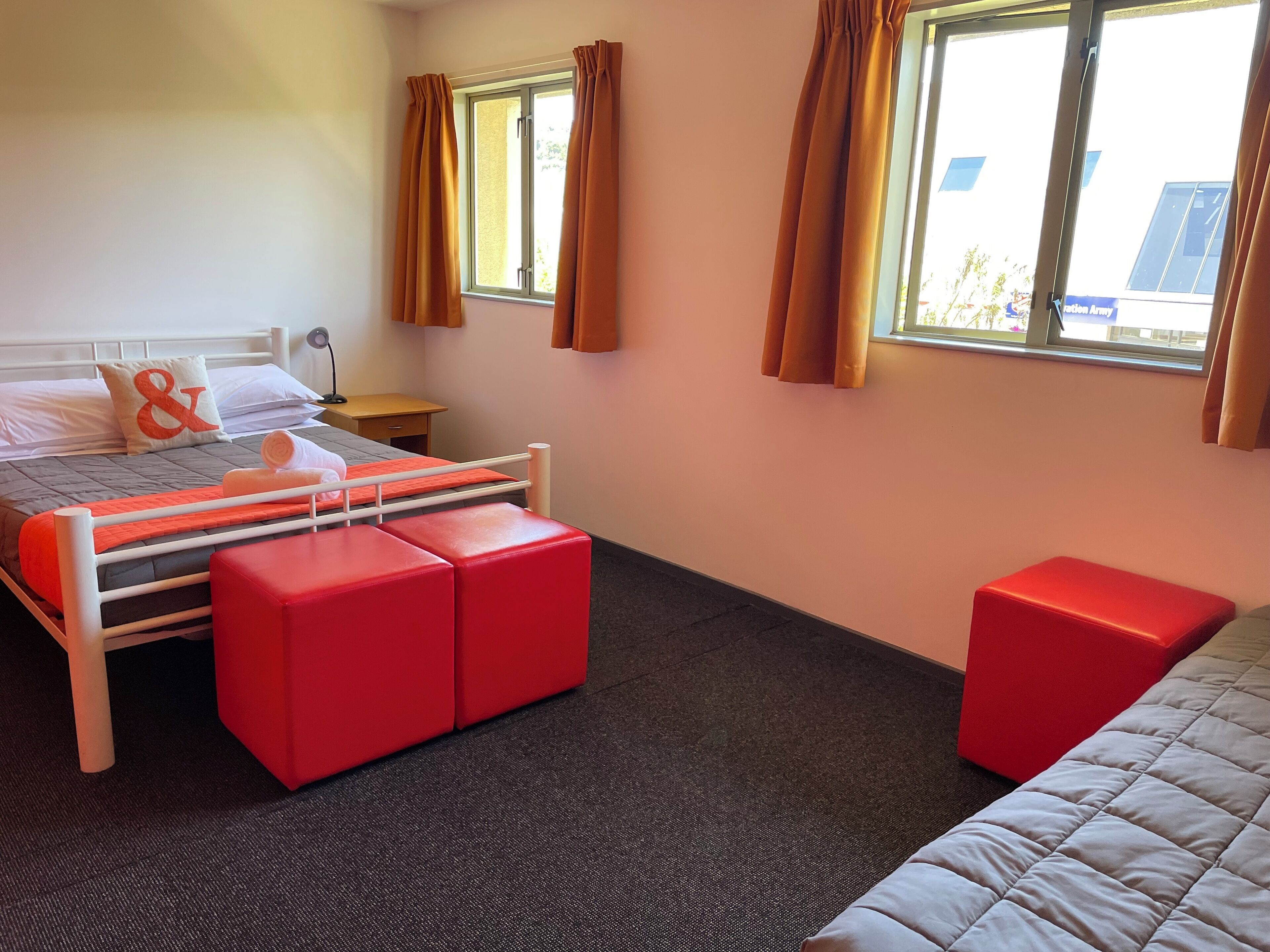 Ett hotellrum med en säng, två röda sittpuffar, ett litet bord och två fönster med gardiner.