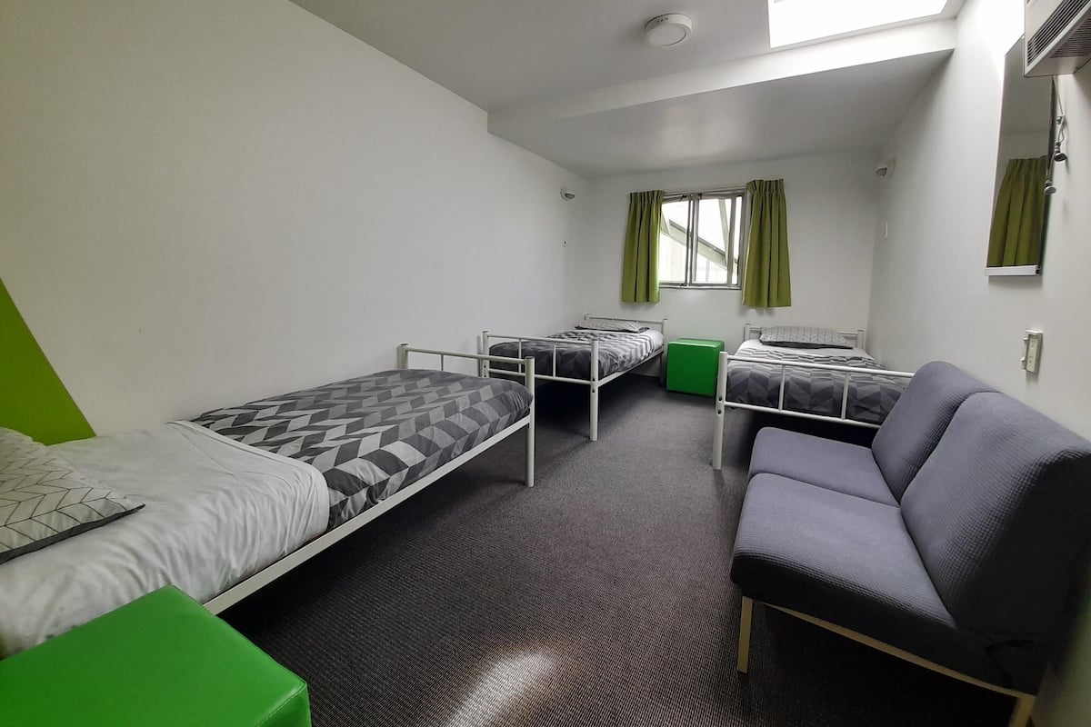 A dormitory room with three single beds, a green storage bench, a grey armchair, and a window with curtains.