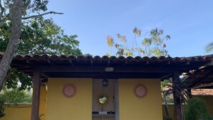 A yellow building with a tiled roof, two circular wall decorations, a small sink, and a black trash bag on the ground. - Bella Geriba Suites (Búzios)