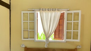 A neatly made bed with white pillows and a grey blanket, a wooden headboard, and a window with open shutters and a curtain. - Bella Geriba Suites (Búzios)