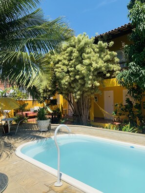 A pool area with a clear blue pool, surrounded by potted plants and a yellow building in the background. - Bella Geriba Suites (Búzios)
