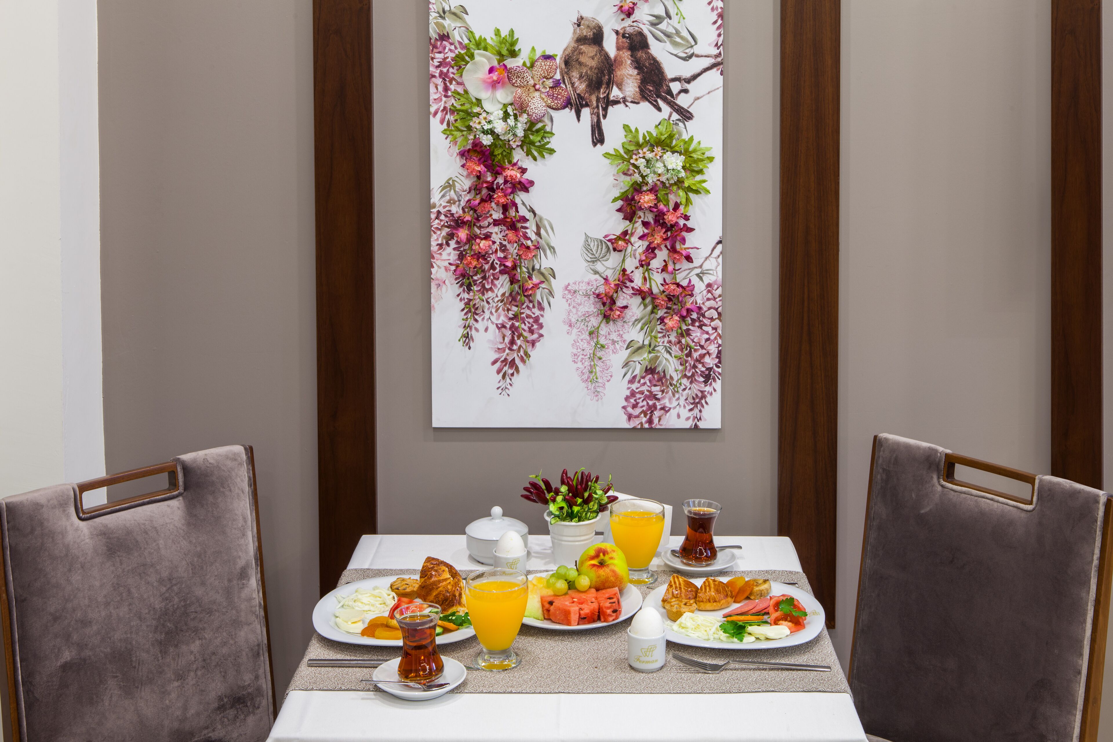Een ontbijttafel met borden eten, glazen jus d'orange en een schilderij van vogels en bloemen aan de muur.