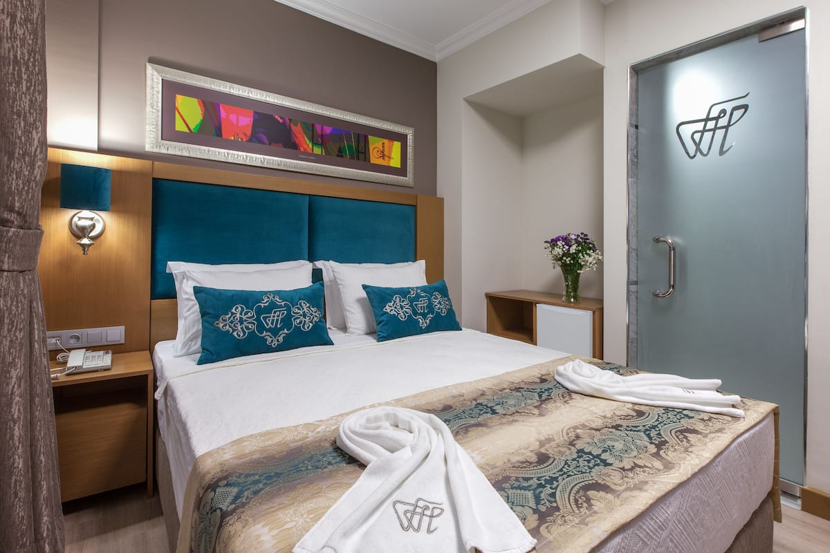 A hotel room with a bed, a nightstand, a wall-mounted phone, a door with a handle, and a framed artwork above the bed.