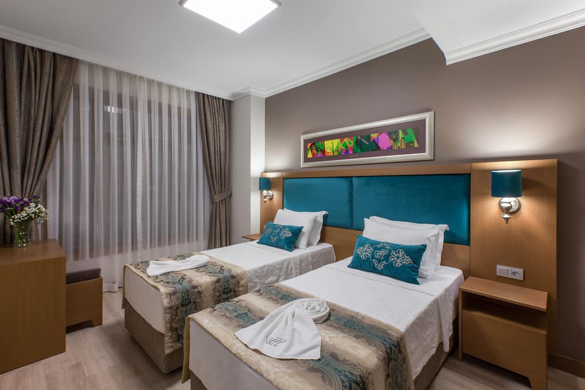 A hotel room with two beds, a large window with curtains, a framed artwork on the wall, and a small table with a vase of flowers.