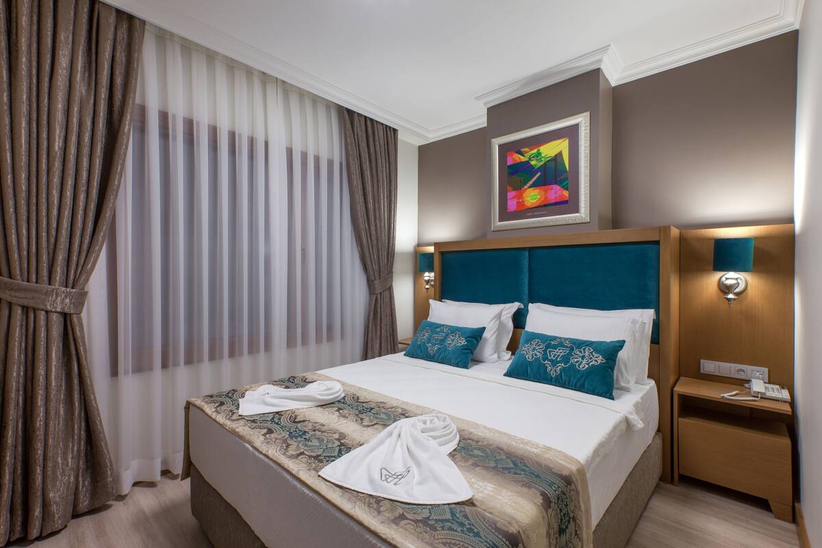A hotel room with a large bed, a bedside table, a wall-mounted light, and a painting above the bed.
