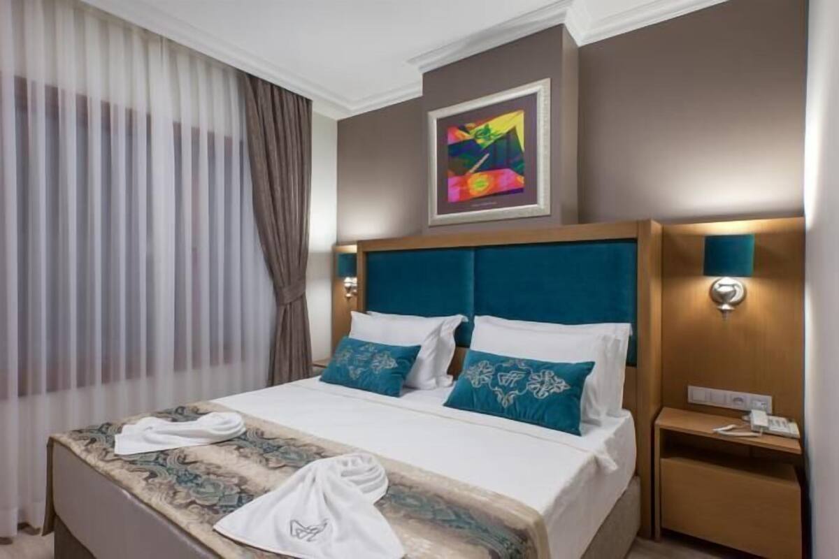A hotel room with a large bed, a headboard, a nightstand, a wall-mounted light, and a painting above the bed.