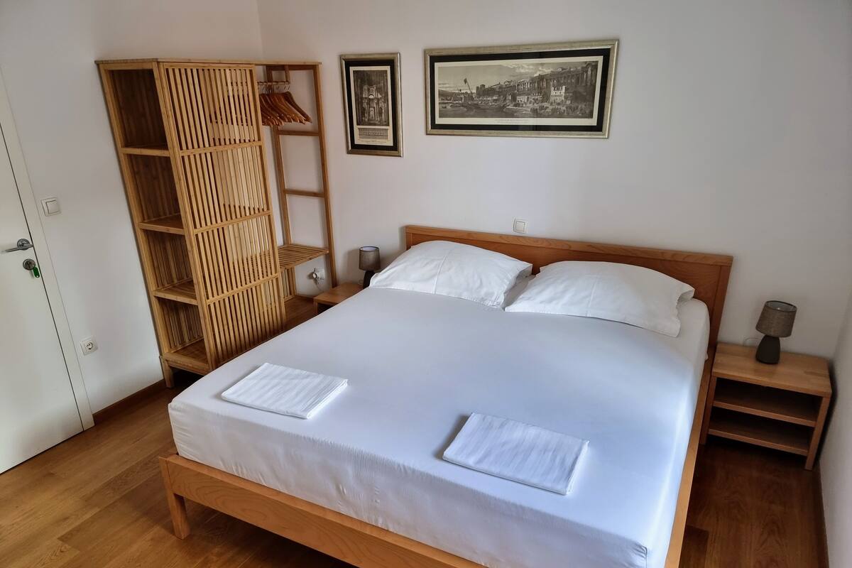 A neatly made bed with white linens and two pillows, a wooden wardrobe with hanging space, a small bedside table with a lamp, and two framed pictures on the wall.
