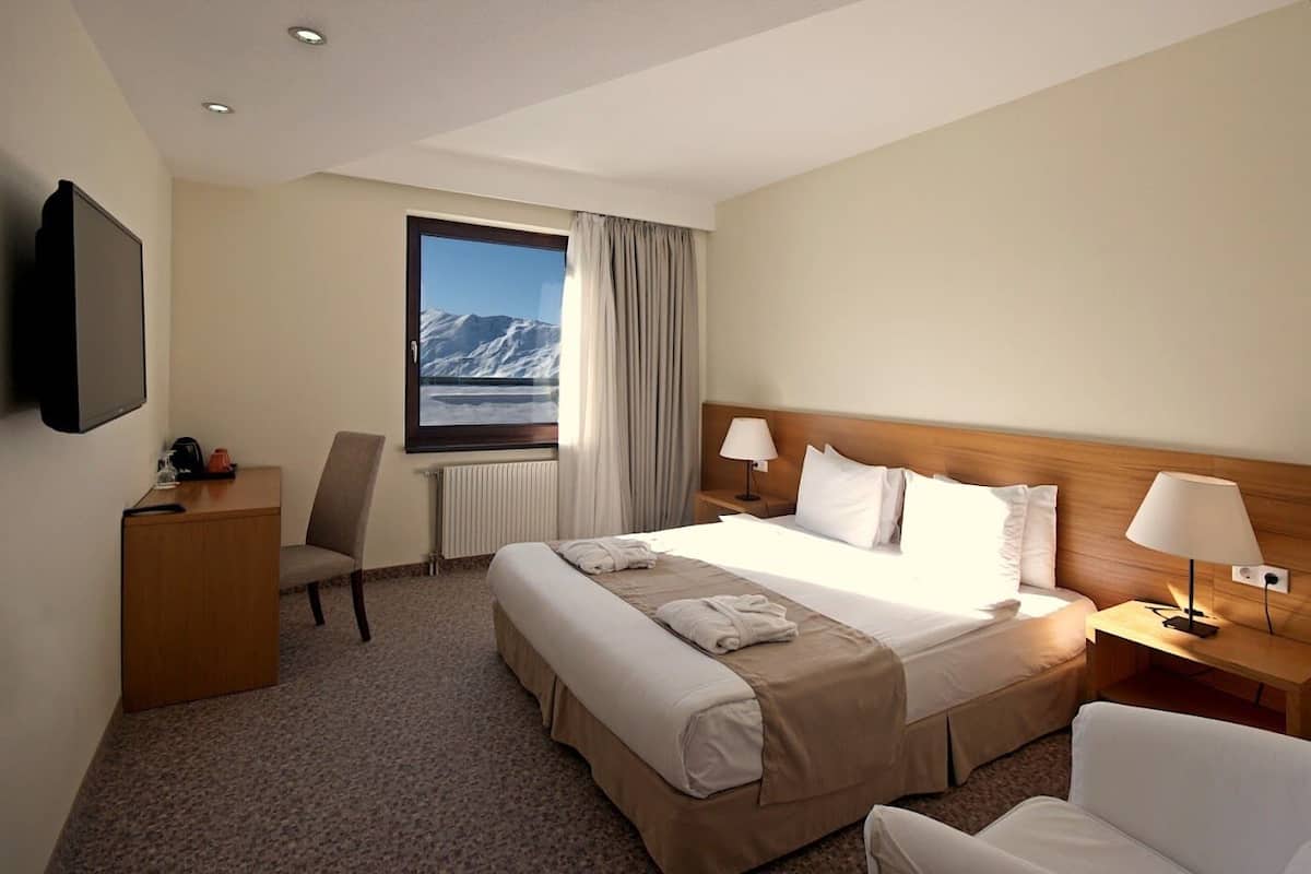 Habitación de hotel con cama, dos mesitas de noche, una silla, escritorio, televisor y ventana con vista a la montaña.