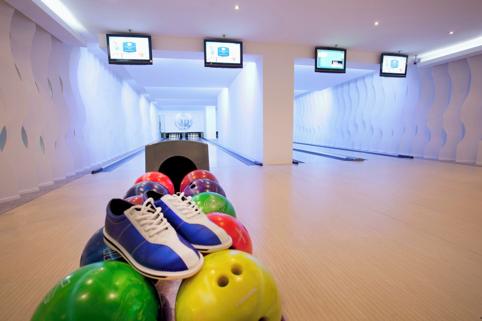 A bowling alley with multiple lanes, a bowling ball rack, and overhead screens displaying advertisements.
