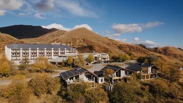 A modern hotel with multiple buildings, including a large structure with balconies and smaller houses with solar panels, set against a backdrop of hills and clear skies.