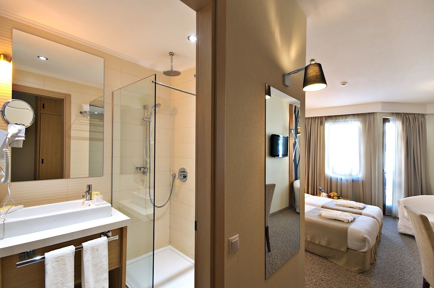 A hotel room with a glass-enclosed shower, a large mirror, a vanity with a sink, and a bed with two pillows.