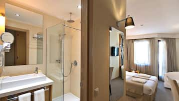 A hotel room with a glass-enclosed shower, a large mirror, a vanity with a sink, and a bed with two pillows.