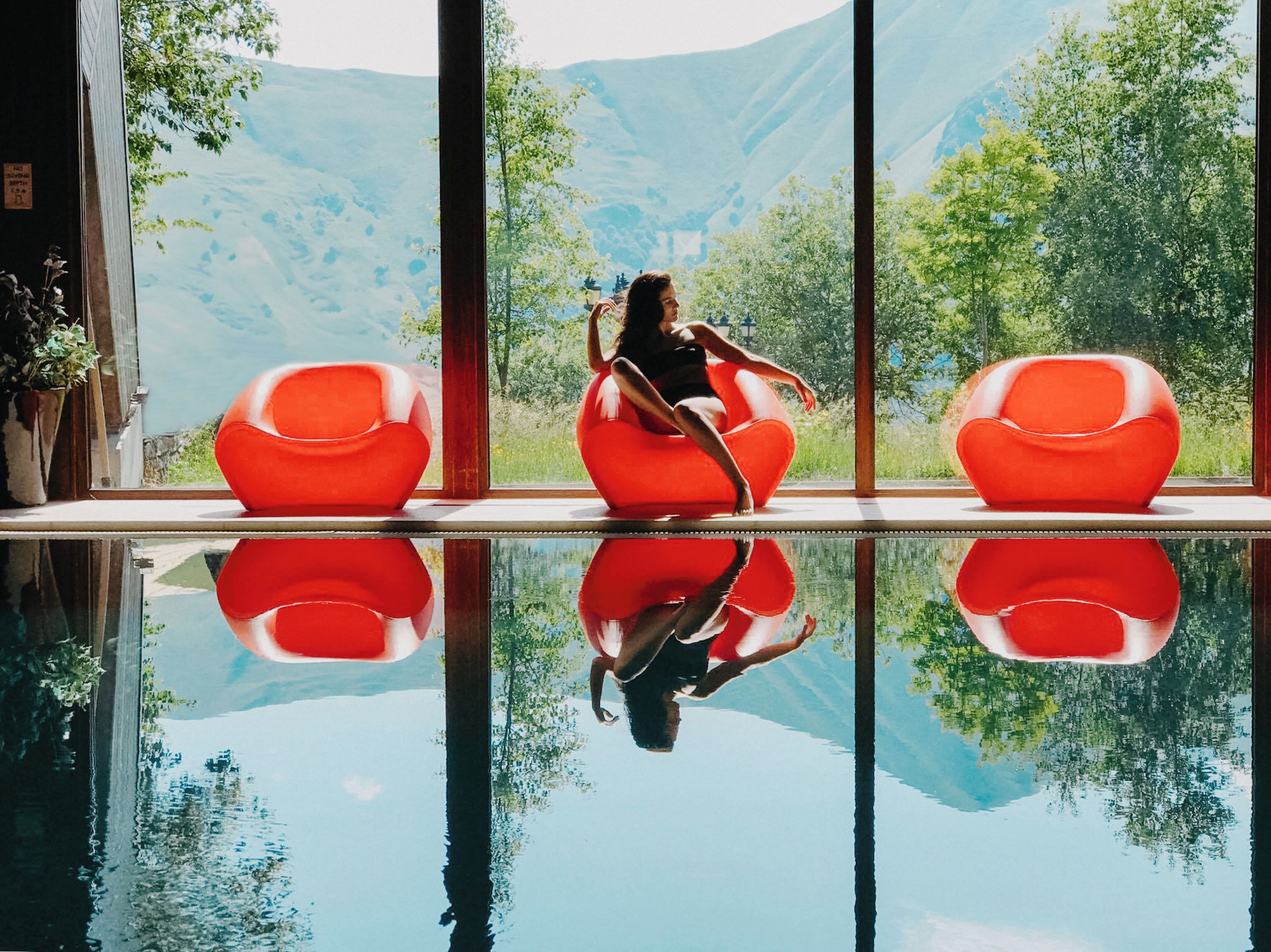 Una persona sentada en una silla roja junto a un ventanal con vistas a montañas y árboles.