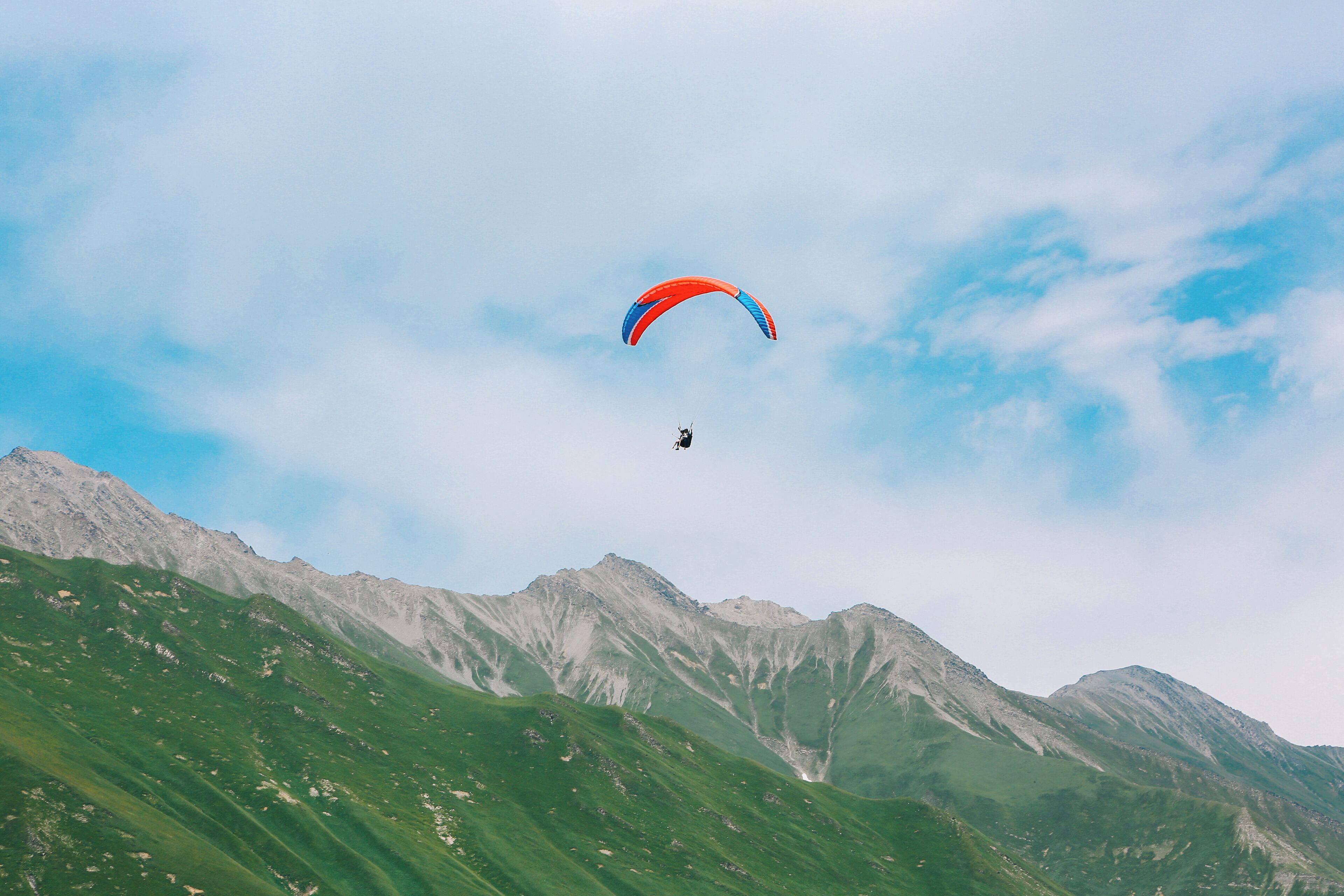 A person paragliding over a mountainous landscape with green slopes and rocky peaks.