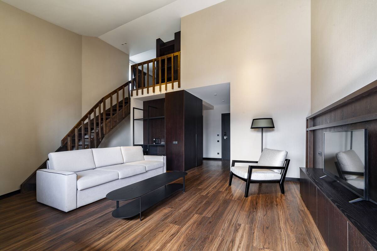 Amplia sala de estar con un sofá blanco, una mesa de centro negra y piso de madera. Una escalera conduce a un nivel superior, y un televisor está montado en la pared.
