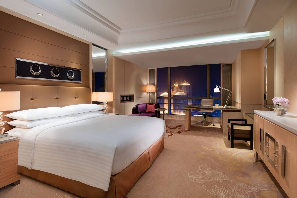 A modern hotel room with a large bed, a desk, and a view of the cityscape.