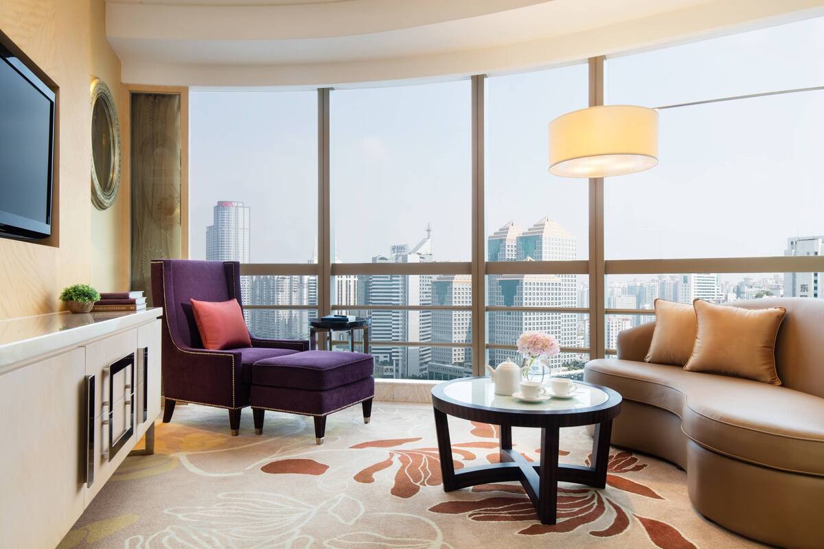 A modern hotel room with a large city view, a purple armchair, a beige sofa, and a round coffee table.