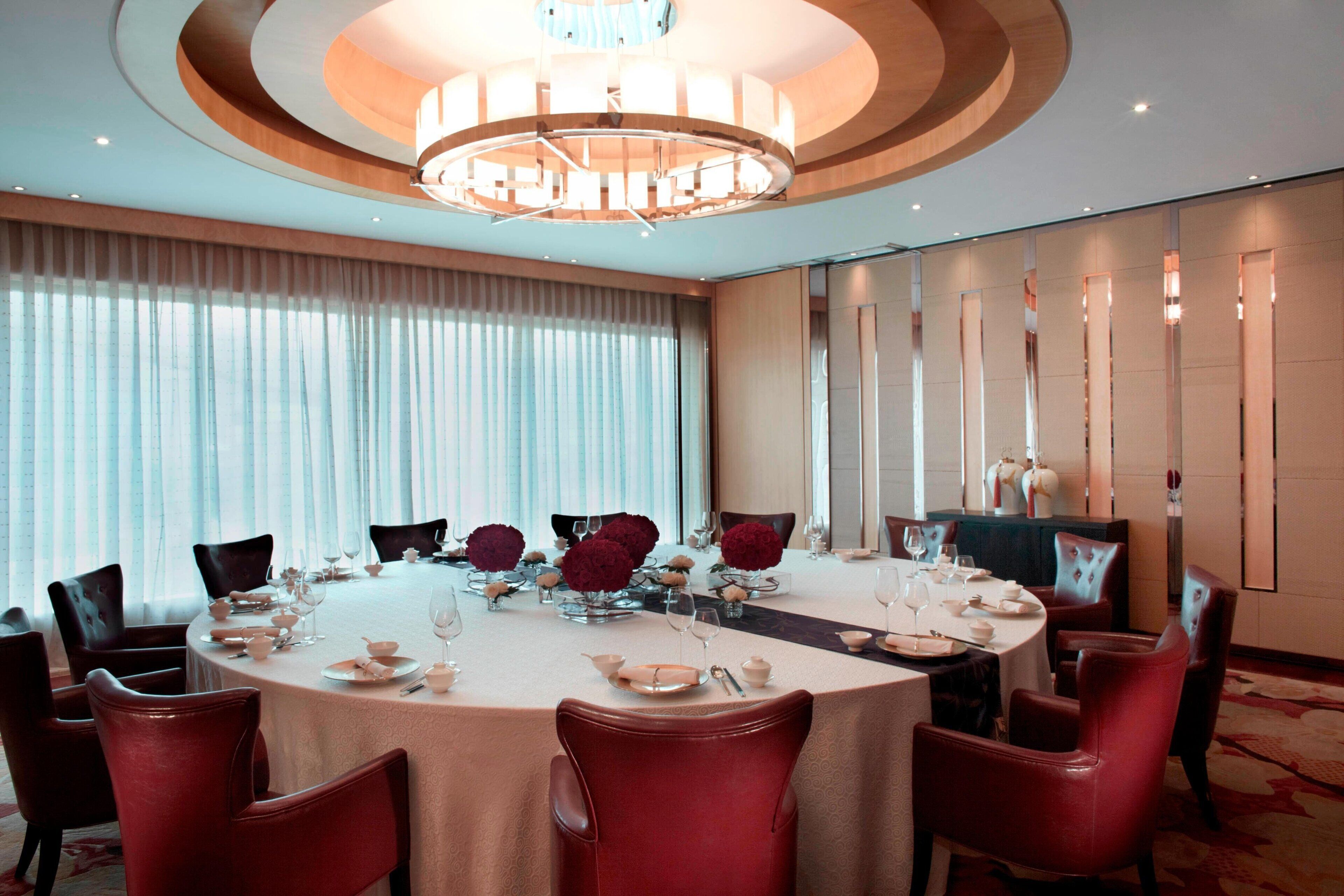 Une table de salle à manger ronde, entourée de chaises rouges, dressée avec de la belle porcelaine et de beaux verres, dans une pièce bien éclairée où trône un grand lustre.