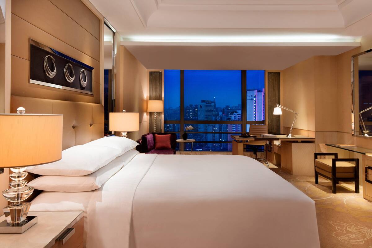 A modern hotel room with a large bed, a desk, and a view of the city at dusk.
