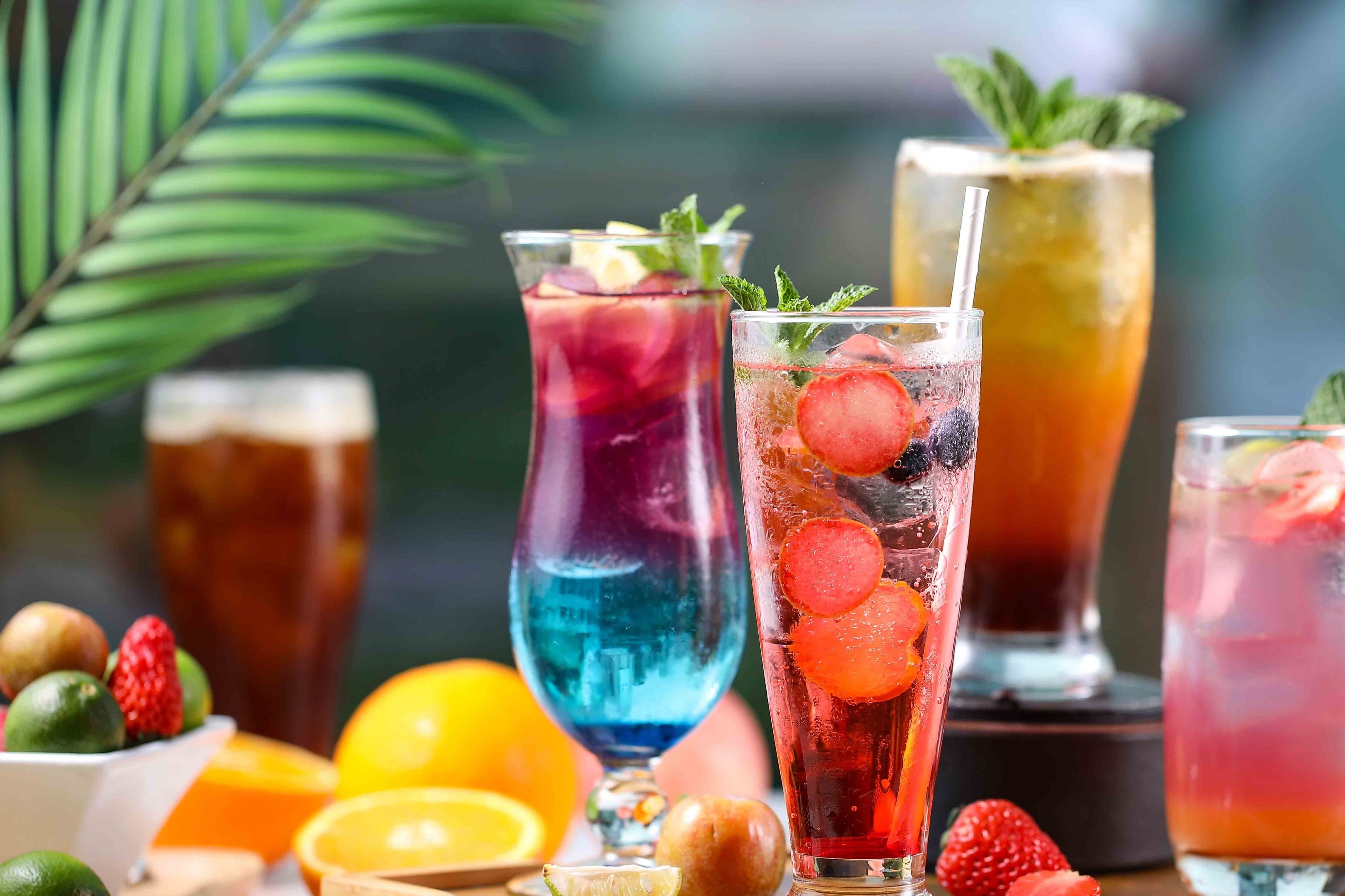 Variedad de bebidas coloridas con frutas y hojas de menta, rodeadas de frutas frescas como fresas y naranjas.