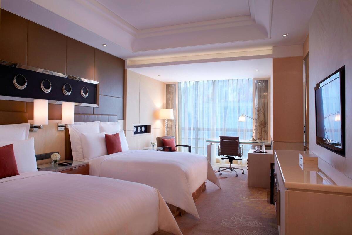 A hotel room with two beds, a desk, a television, and a large window with curtains.