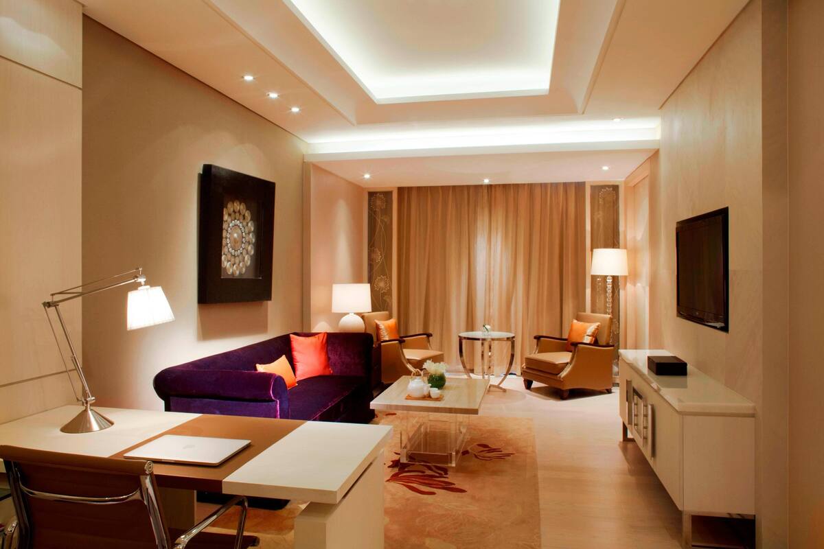 A modern hotel room with a sofa, a dining table, a desk, a TV, and a lamp.