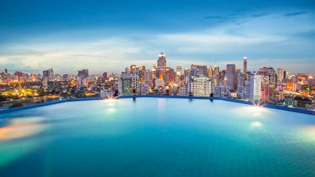 A city skyline at dusk with a large, clear pool in the foreground.