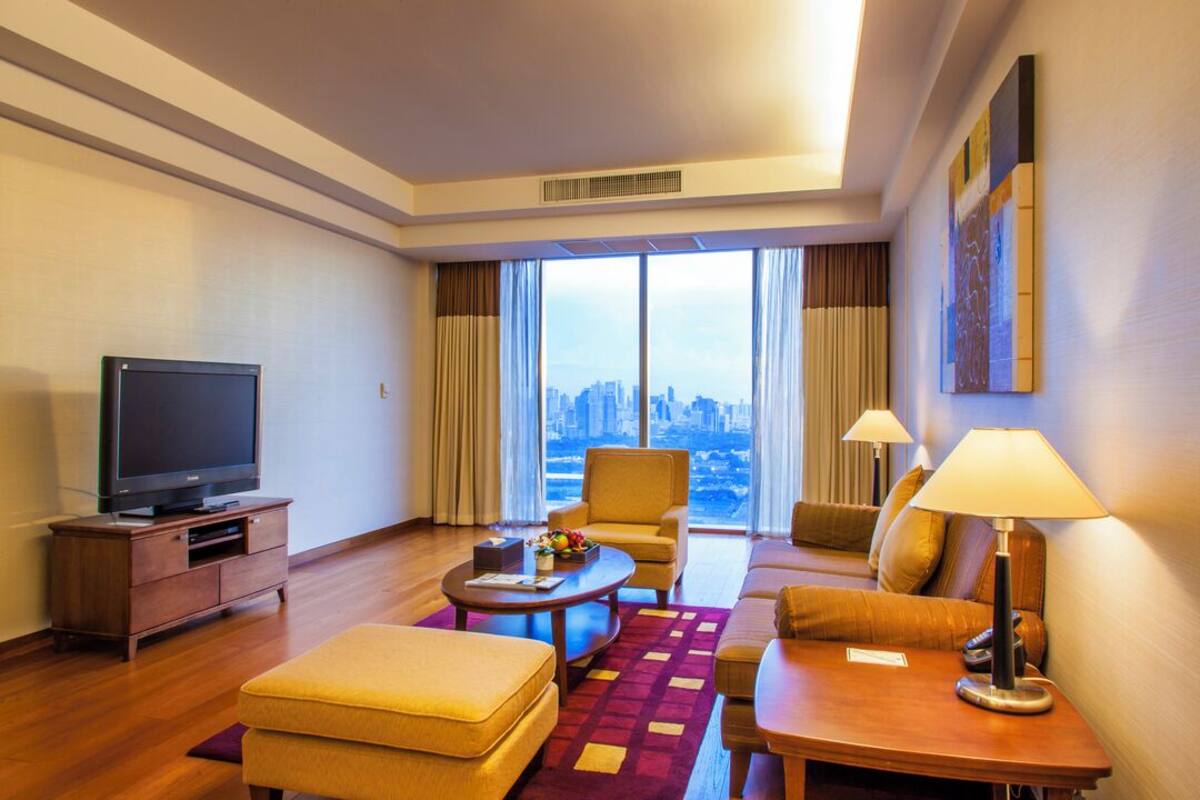 Una habitación de hotel con un ventanal grande, un televisor de pantalla plana, un sofá, una mesita de centro y una lámpara.