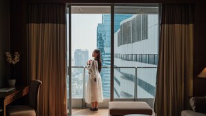 A modern hotel room with a large sliding glass door leading to a balcony, a wooden desk, a cushioned chair, and a round table.