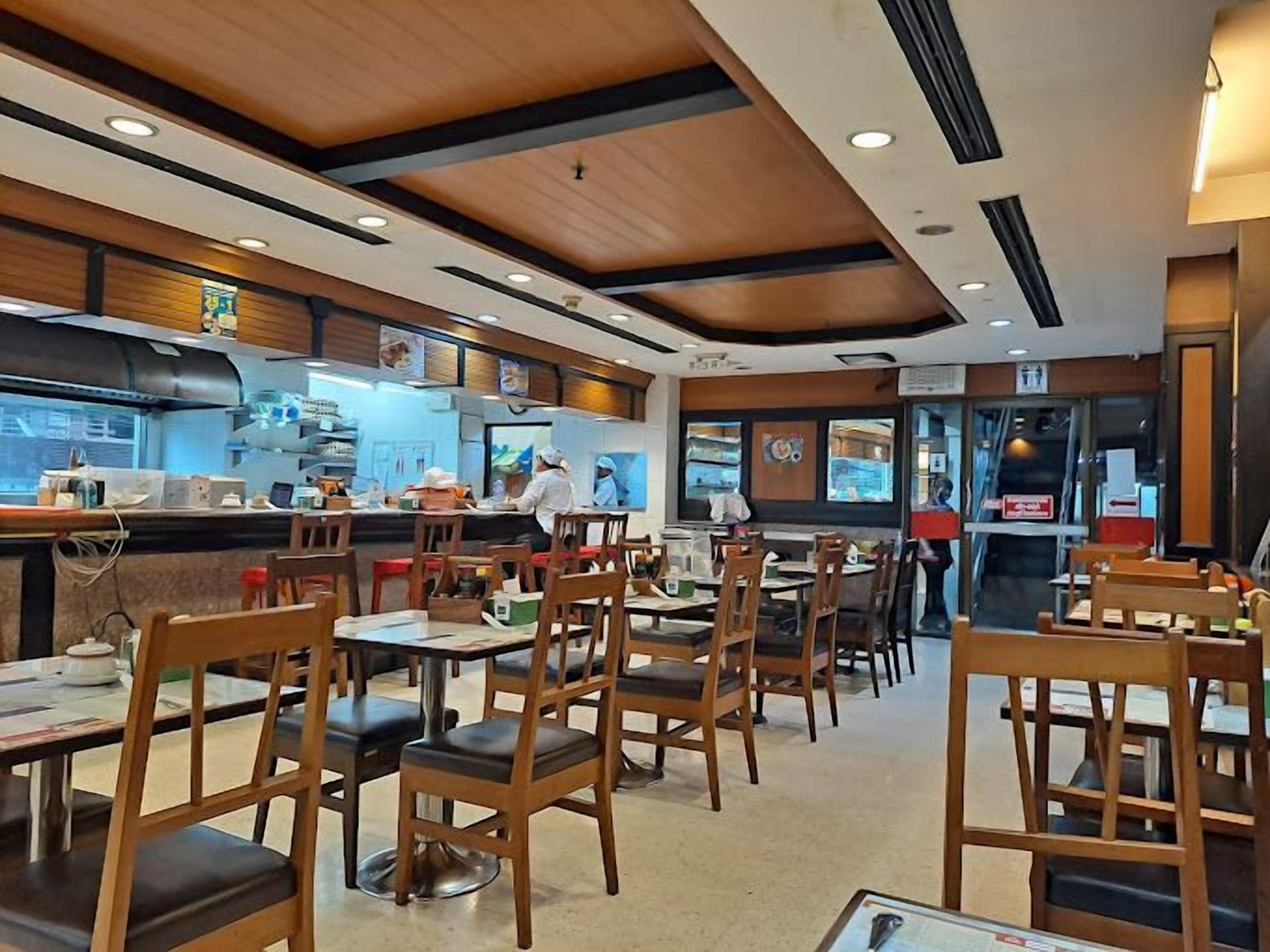 A restaurant interior with wooden tables and chairs, a kitchen area, and a counter where staff are preparing food.