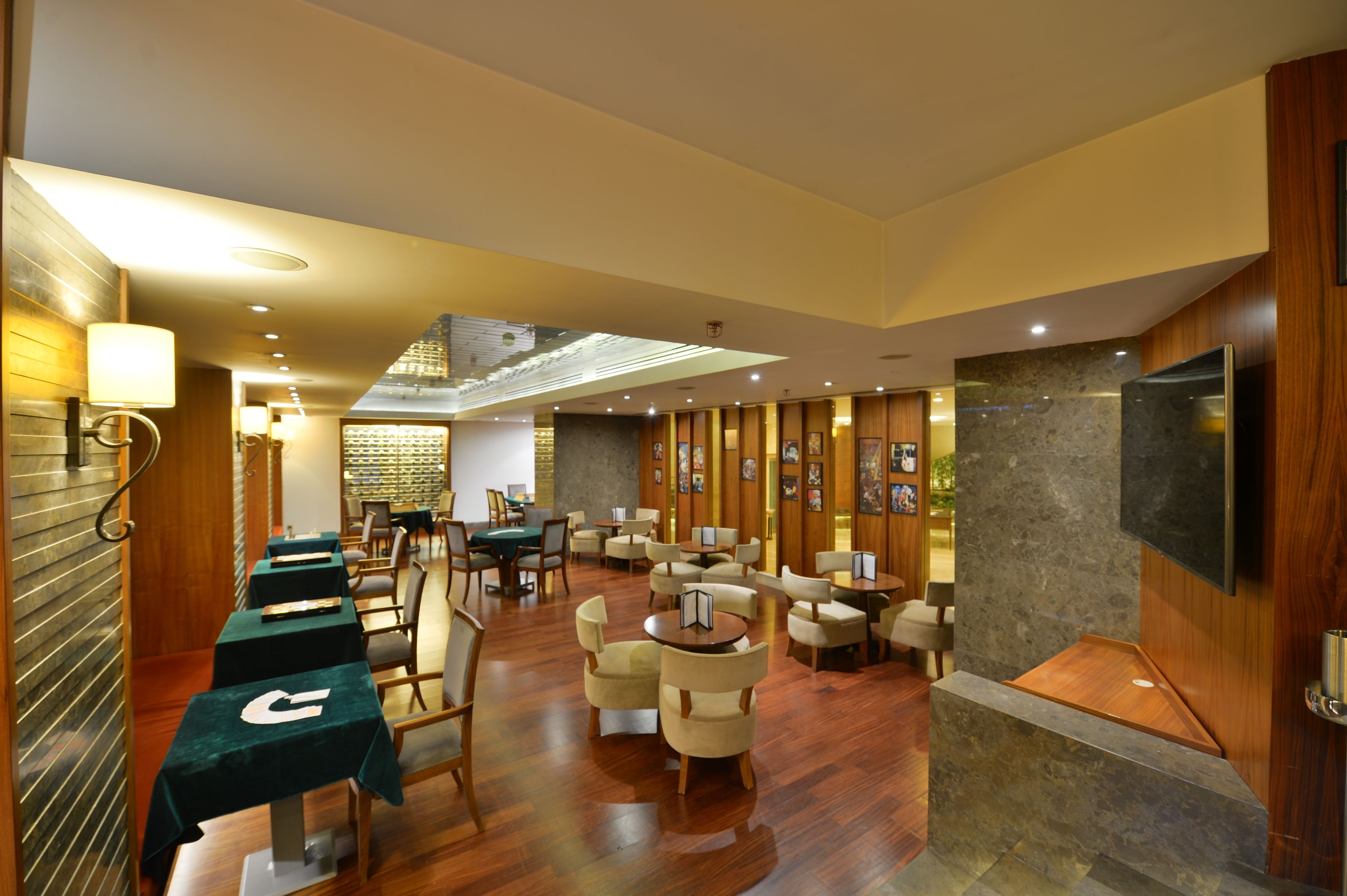 Un hall d’hôtel moderne avec un plancher en bois, des tables vertes et des chaises beiges. On y trouve des téléviseurs fixés au mur et un grand mur décoratif en pierre.