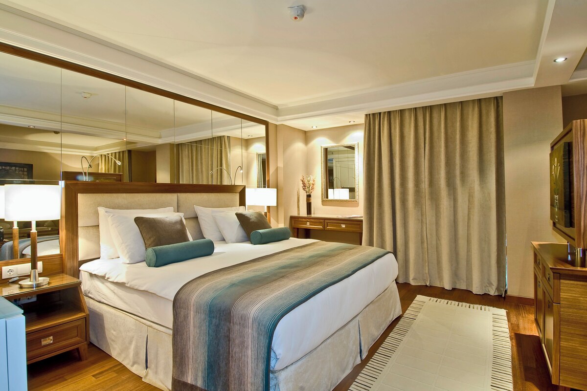 A hotel room with a large bed, a nightstand with a lamp, a dresser, a mirror, and a television.