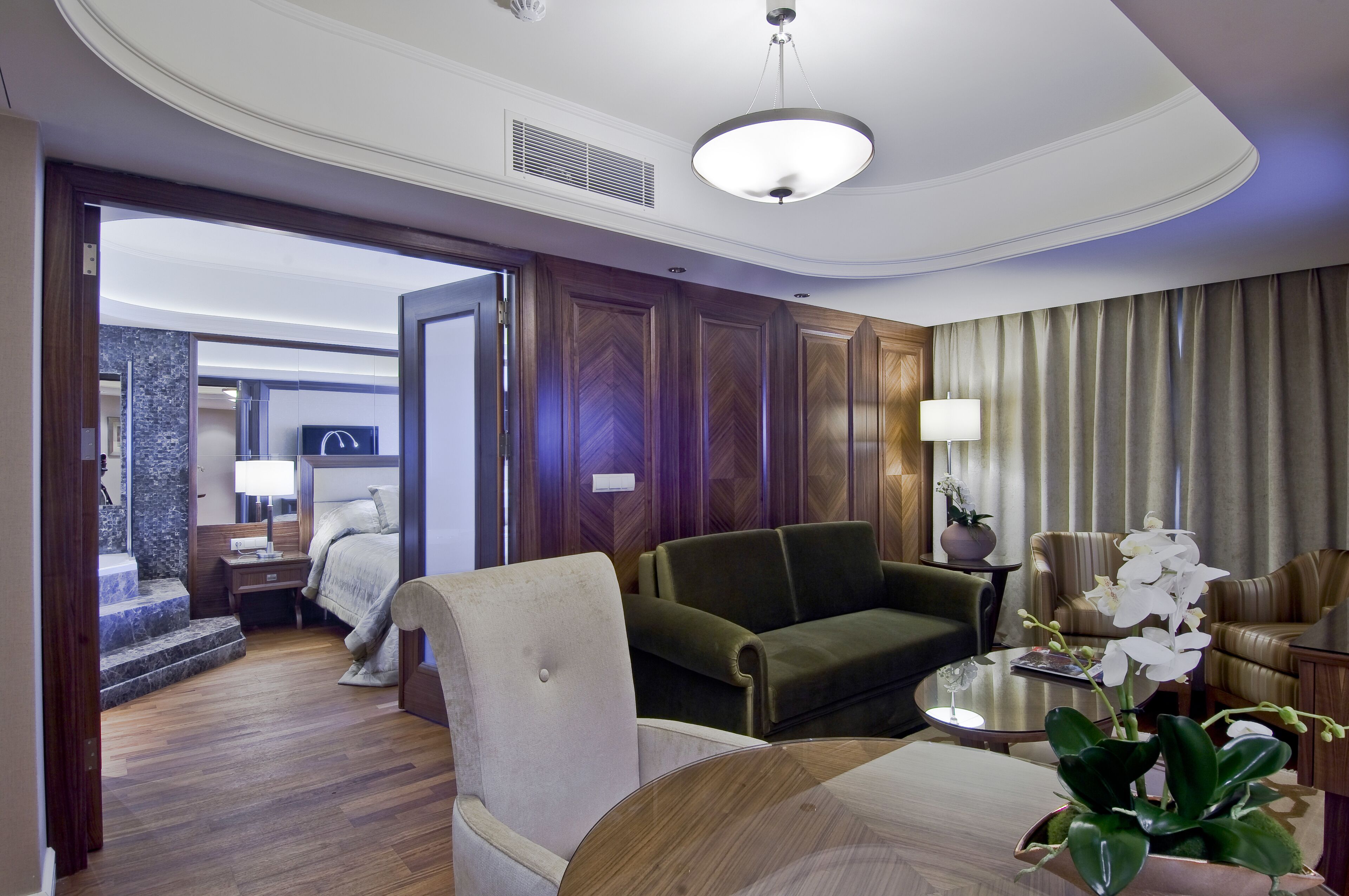 A hotel room with a wooden floor, a green sofa, a white armchair, a round coffee table, and a potted orchid.