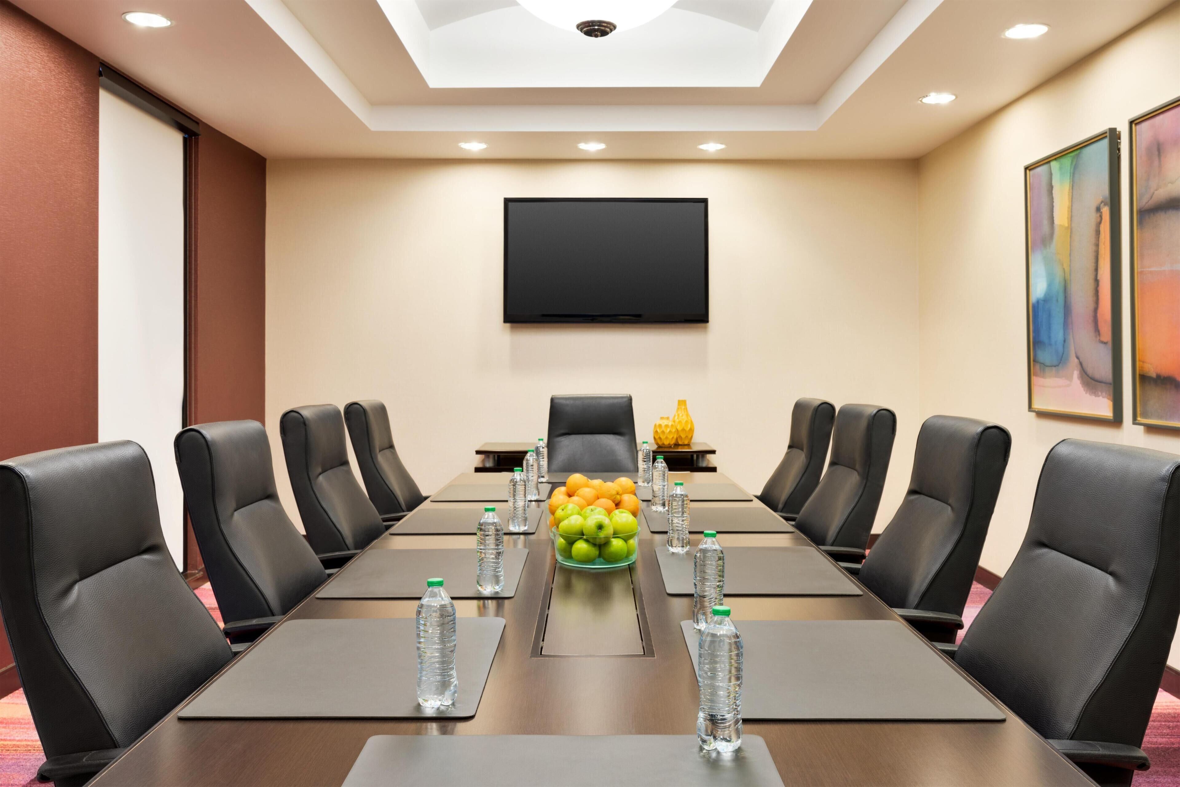 Un salón de conferencias con una mesa larga, sillas de cuero negro, un televisor de pantalla plana y un frutero en el centro.