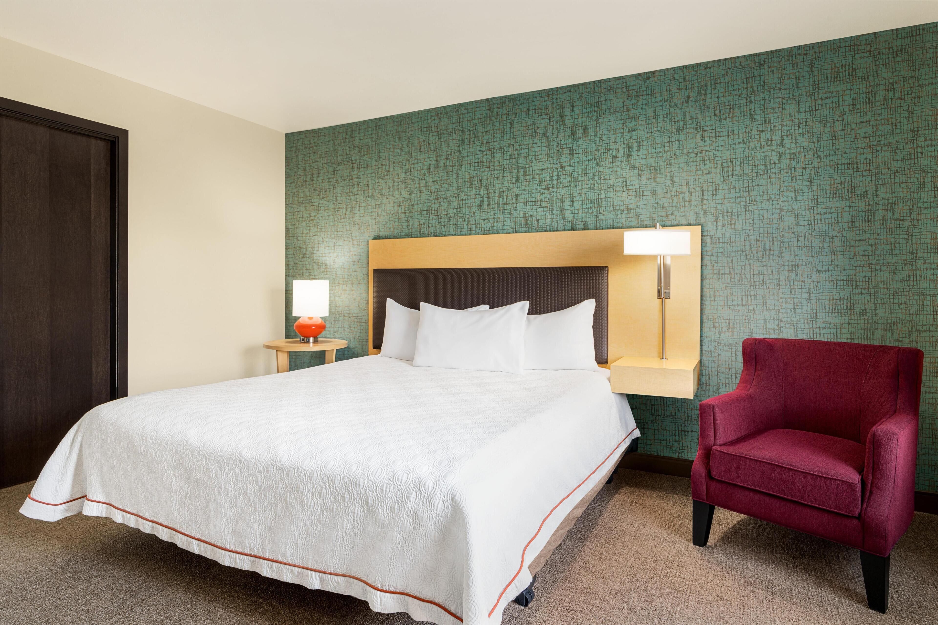 A hotel room with a large bed, a red armchair, a nightstand with a lamp, and a green textured wall.