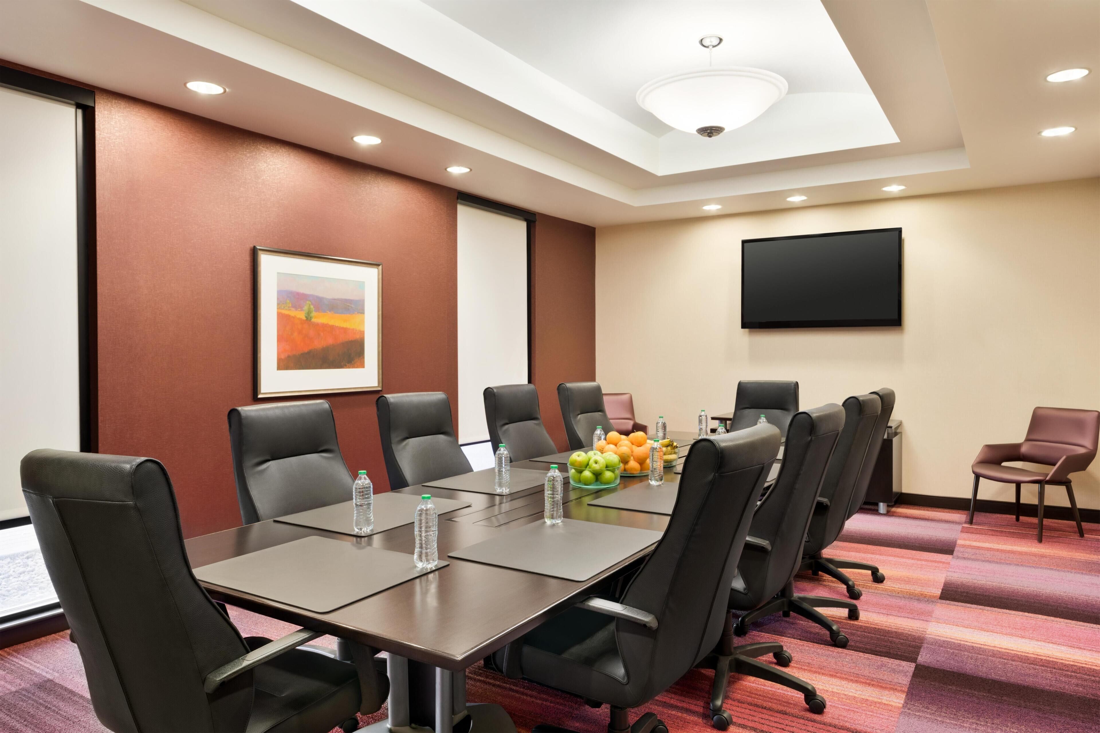 Un salón de conferencias con una larga mesa, varias sillas, una pantalla de proyector y un frutero.