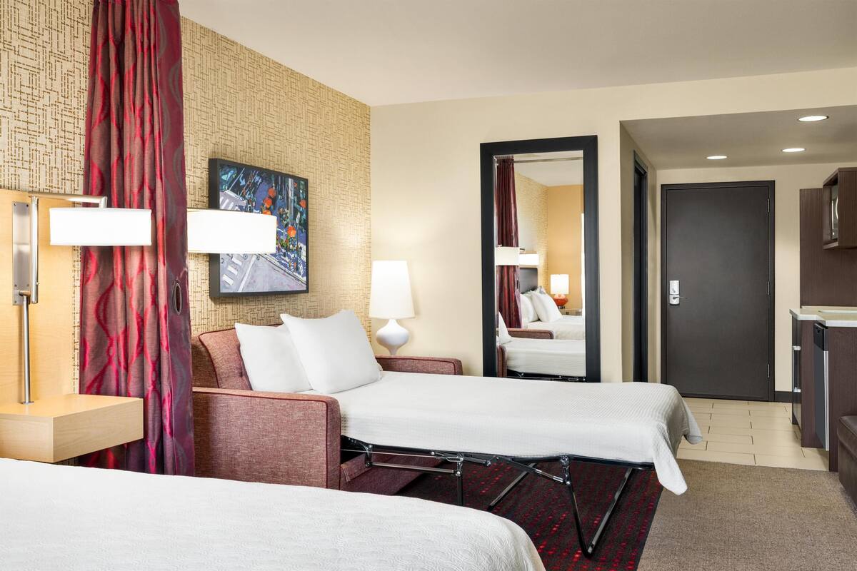 Habitación de hotel con dos camas, un espejo grande, un cuadro en la pared y un baño visible a través de una puerta abierta.