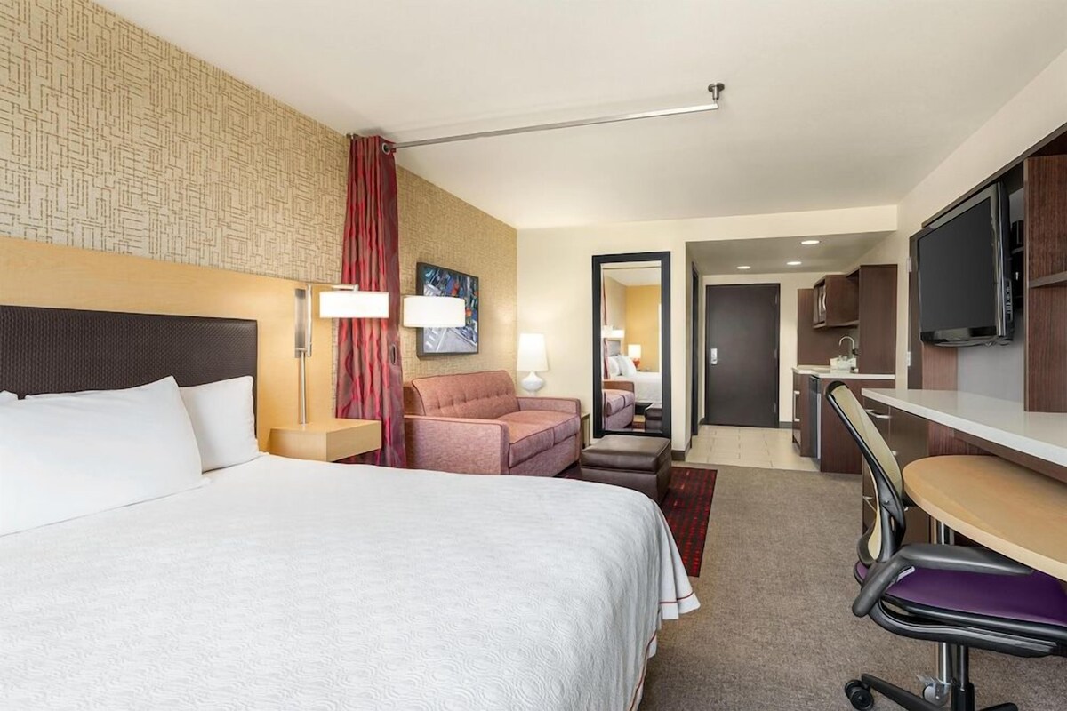 A hotel room with a bed, a desk, a chair, a TV, a mirror, and a door leading to a bathroom.