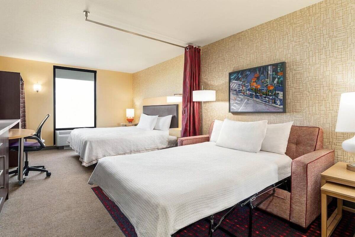 Habitación de hotel con dos camas, un escritorio, una silla, un televisor, un cuadro y una ventana.