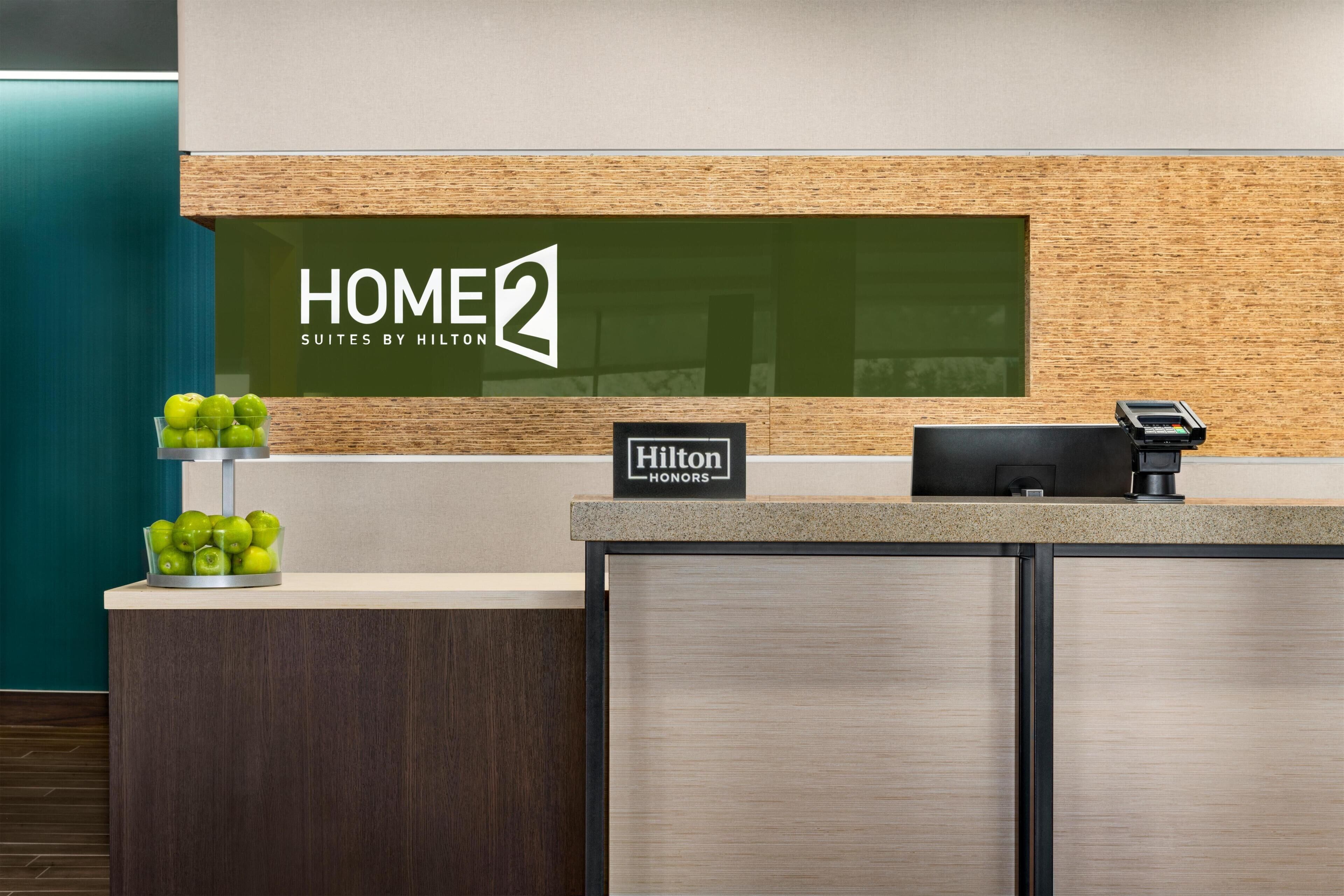 A reception desk with a "HOME2 Suites by Hilton" sign, a "Hilton Honors" badge, a digital payment terminal, and a bowl of green apples.