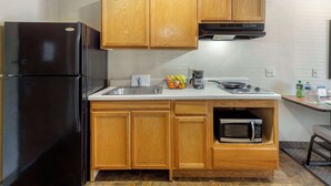 A compact kitchen with wooden cabinets, a black refrigerator, a microwave, and a sink. - WoodSpring Suites Raleigh Garner (Garner)
