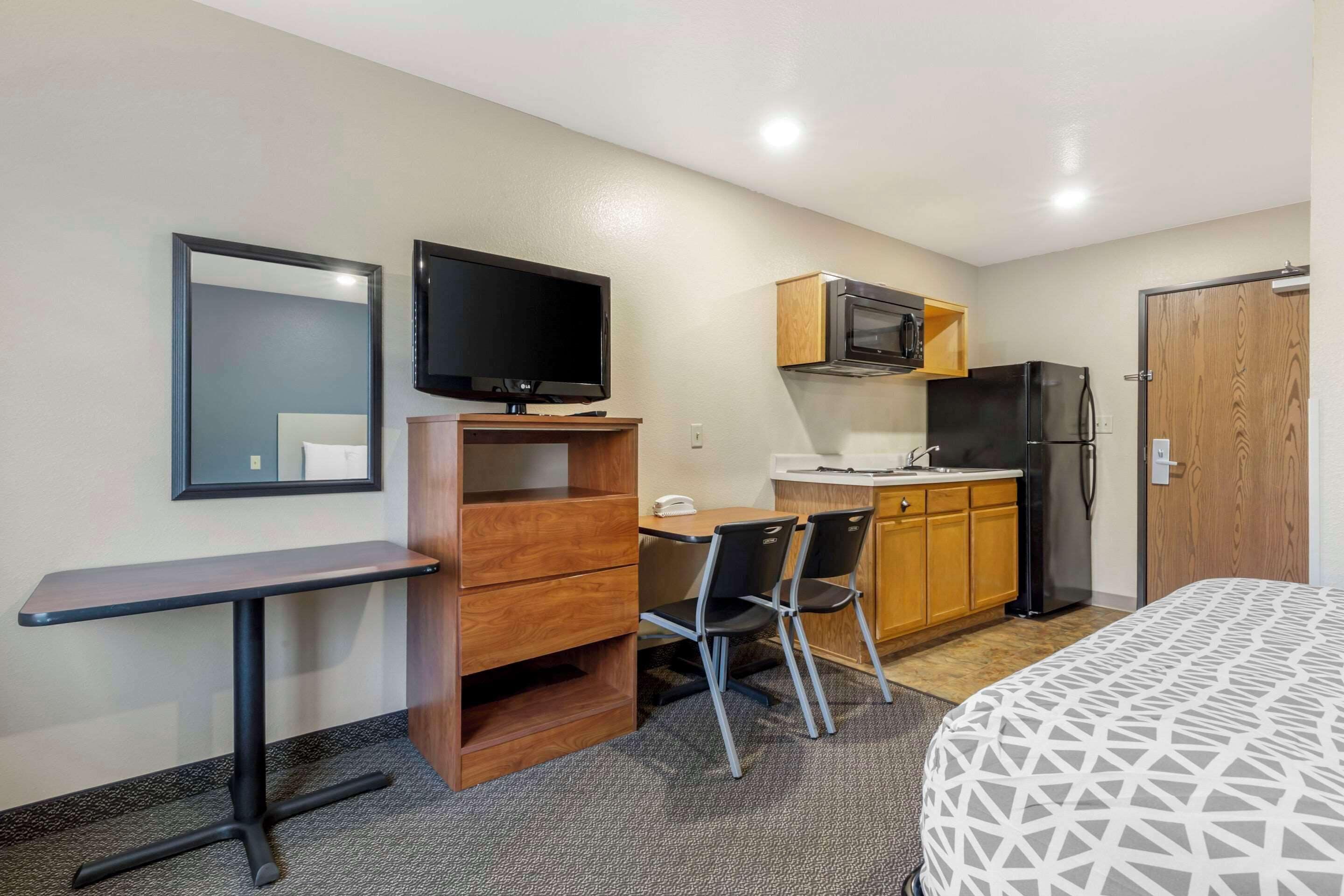 A compact hotel room with a kitchenette, a bed, a desk with a chair, a television, and a mirror.