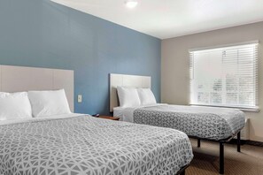 A hotel room with two beds, a large window with blinds, and a blue accent wall. - WoodSpring Suites Raleigh Garner (Garner)
