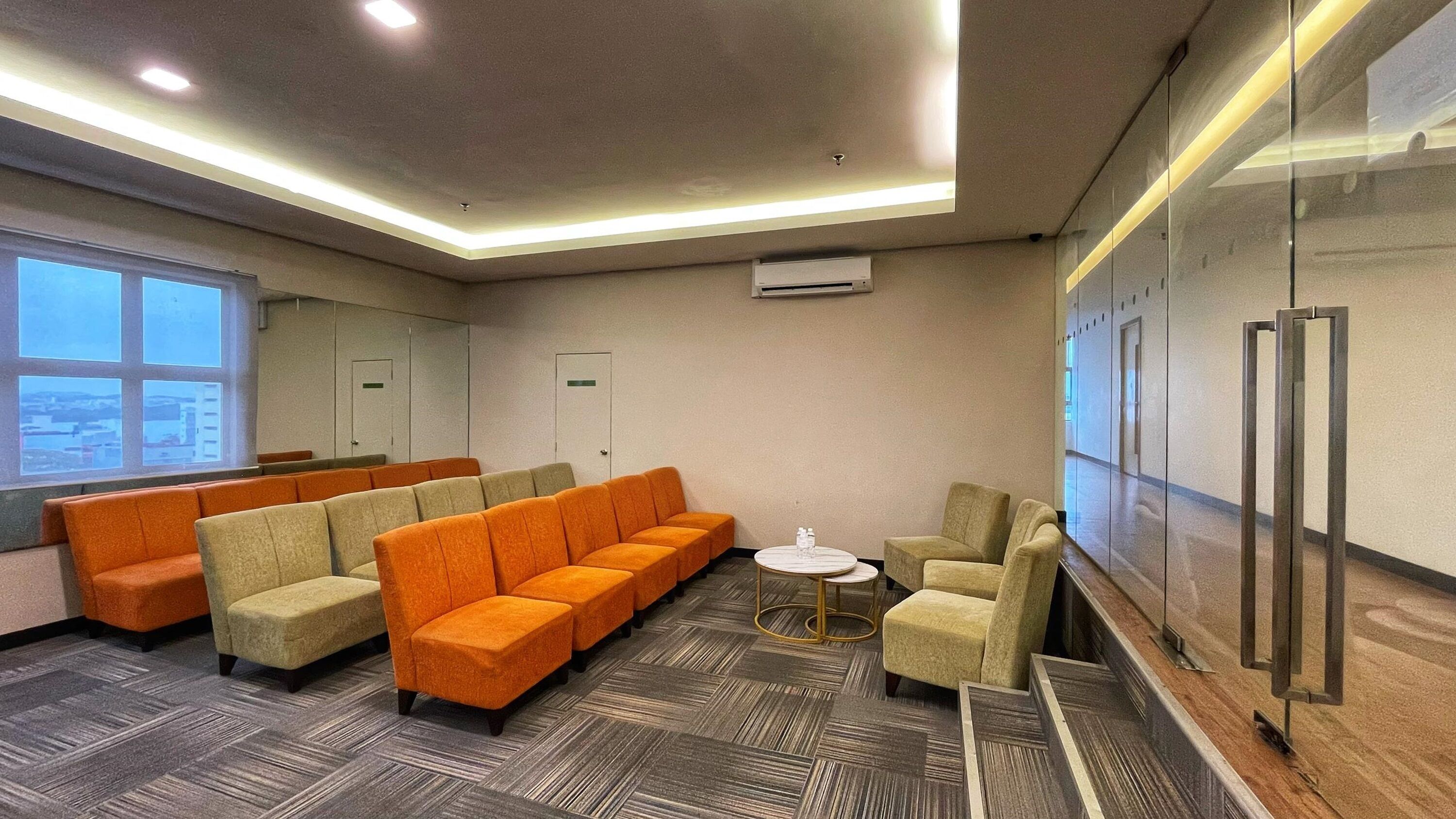 Una sala de conferencias con un sofá naranja largo, un sofá beige más pequeño, una mesa redonda con dos botellas de agua y un muro divisorio de vidrio.