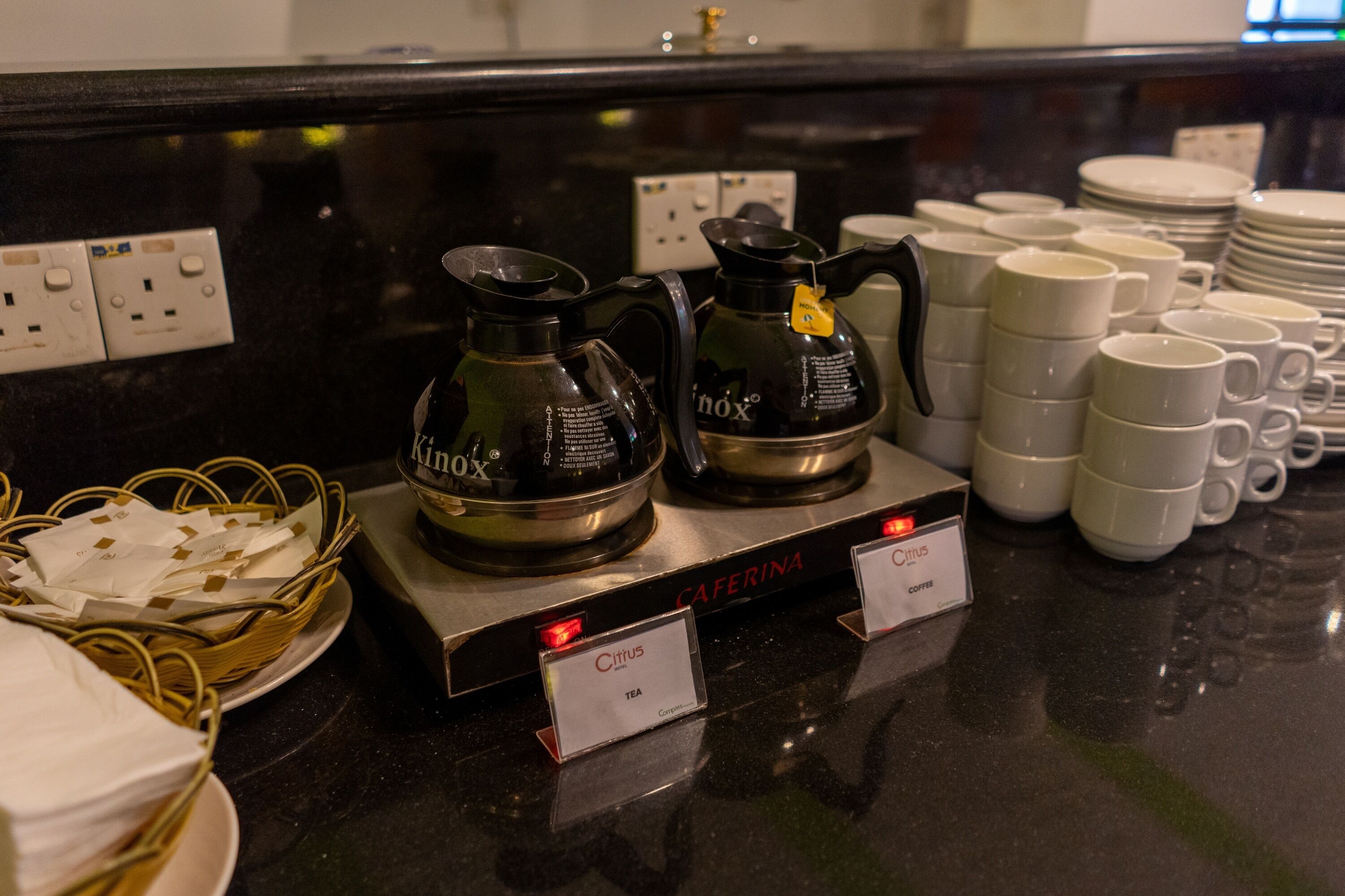 A coffee station with two coffee makers labeled 'Kinox' and 'Cafarina', a basket of napkins, and stacks of white cups and saucers.