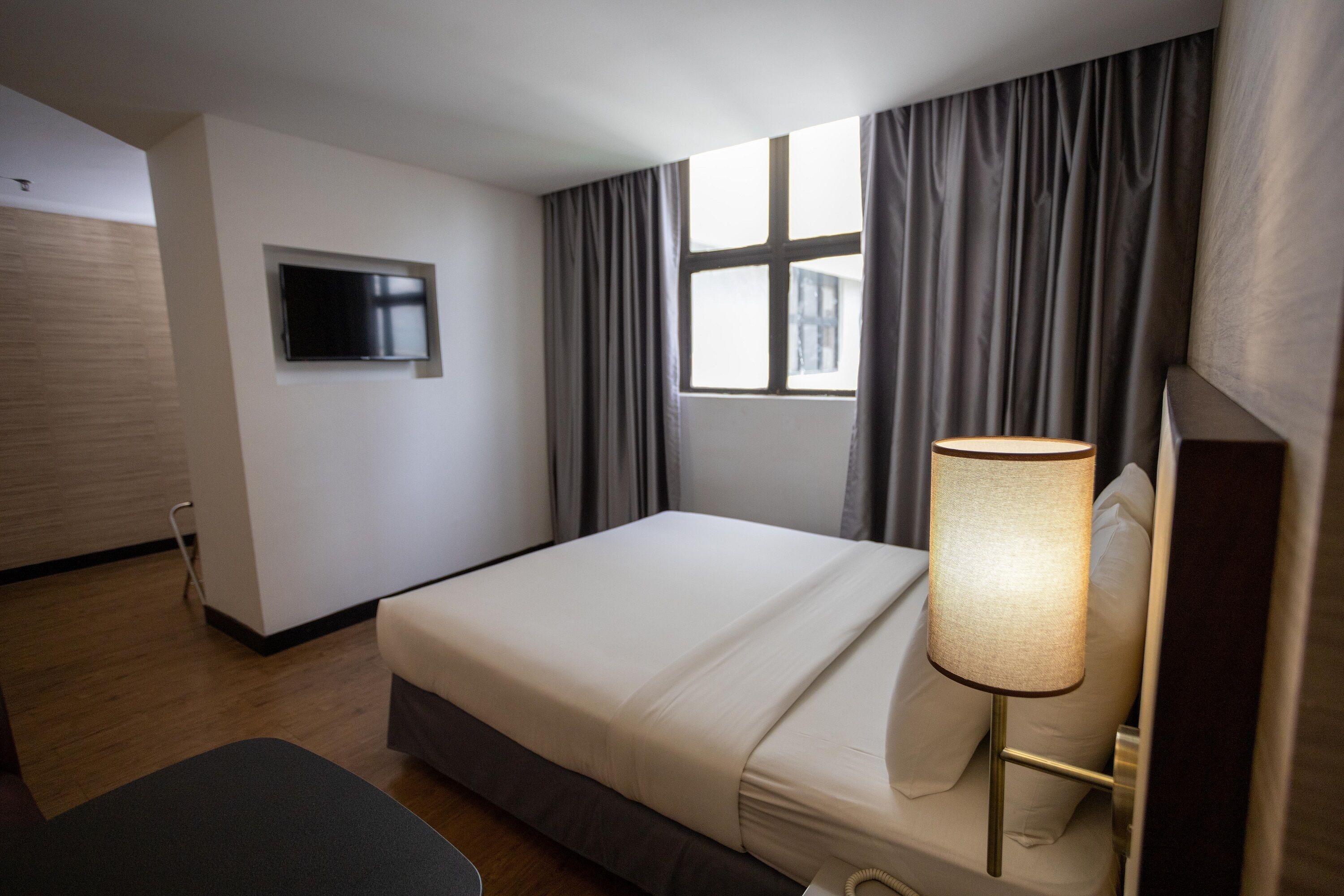 A hotel room with a bed, a TV mounted on the wall, a window with curtains, and a bedside lamp.