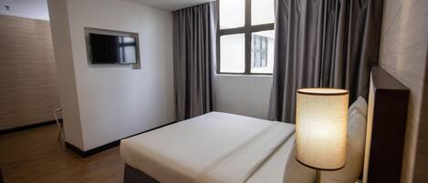 A hotel room with a bed, a TV mounted on the wall, a window with curtains, and a bedside lamp.