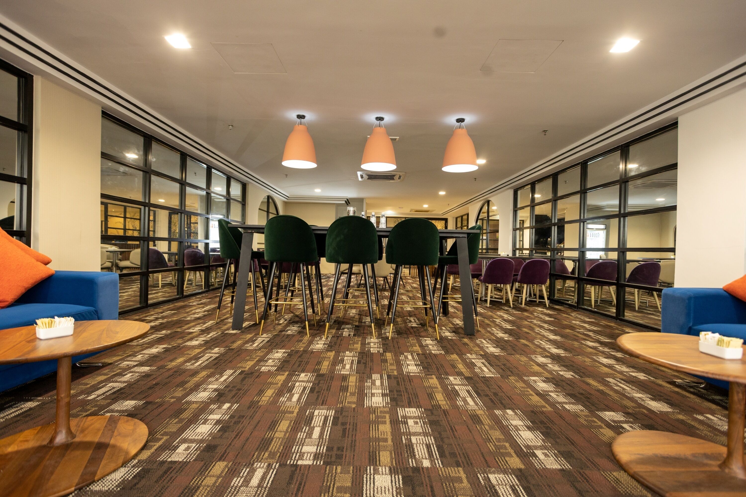 Un moderno vestíbulo de hotel con alfombra estampada, sillas verdes y púrpuras, lámparas colgantes naranjas y sofás azules.