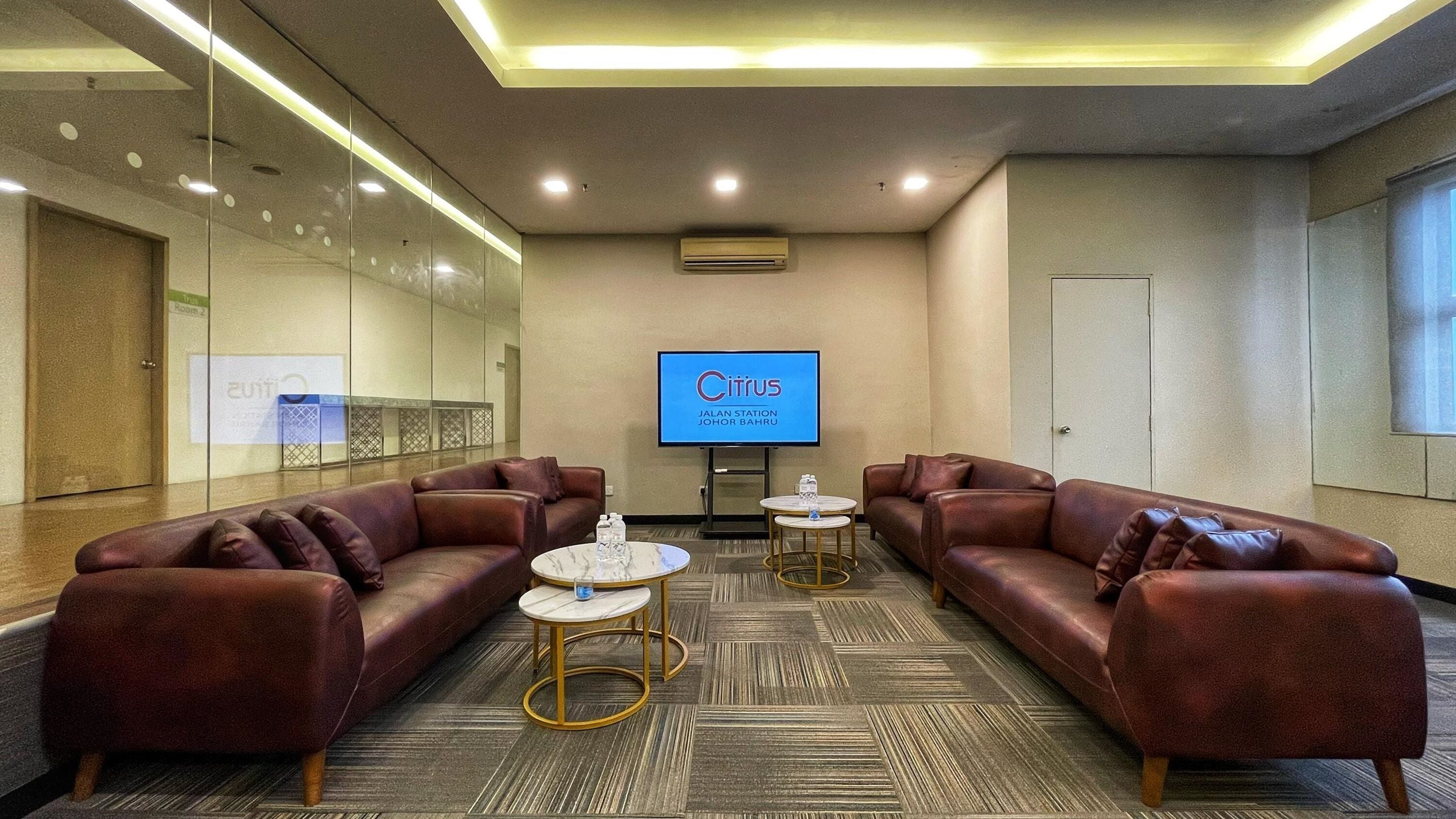 A conference room with leather sofas, small round tables, and a large screen displaying 'Citrus JALAN STATION JOHOR BAHRU'.
