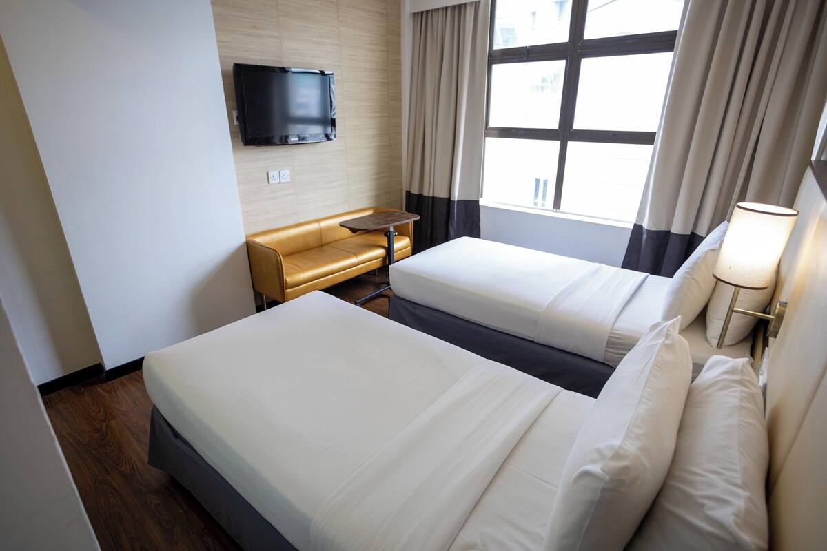 A hotel room with two beds, a TV mounted on the wall, a window with curtains, and a small table.